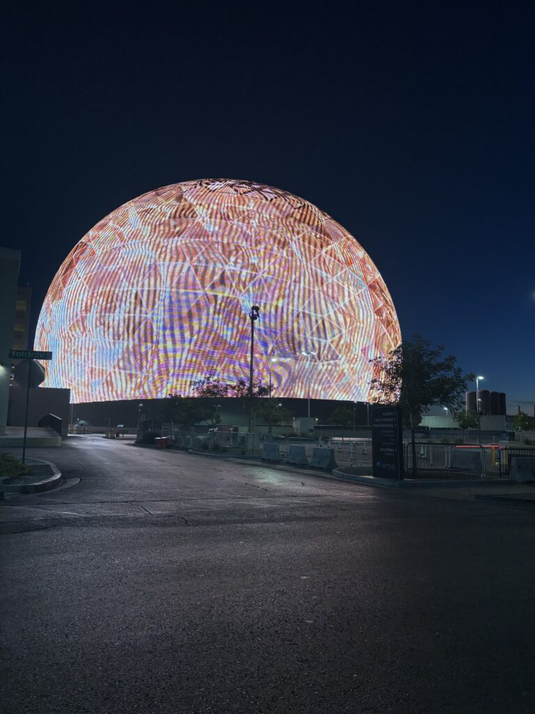 Las Vegas Sphere lit up at dawn, seen from Westchester Drive with signs for parking, valet and rideshare