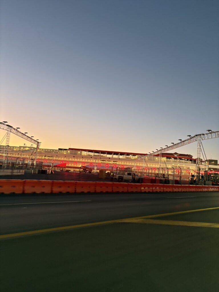 Las Vegas Grand Prix pit building at sunrise, with track lighting rigs and orange barriers along the road