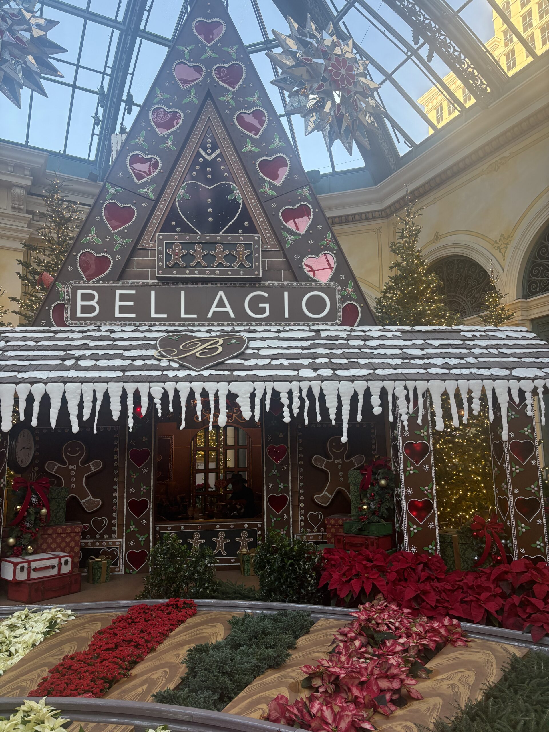 Giant gingerbread house with a BELLAGIO sign surrounded by poinsettias in the Bellagio Conservatory holiday display