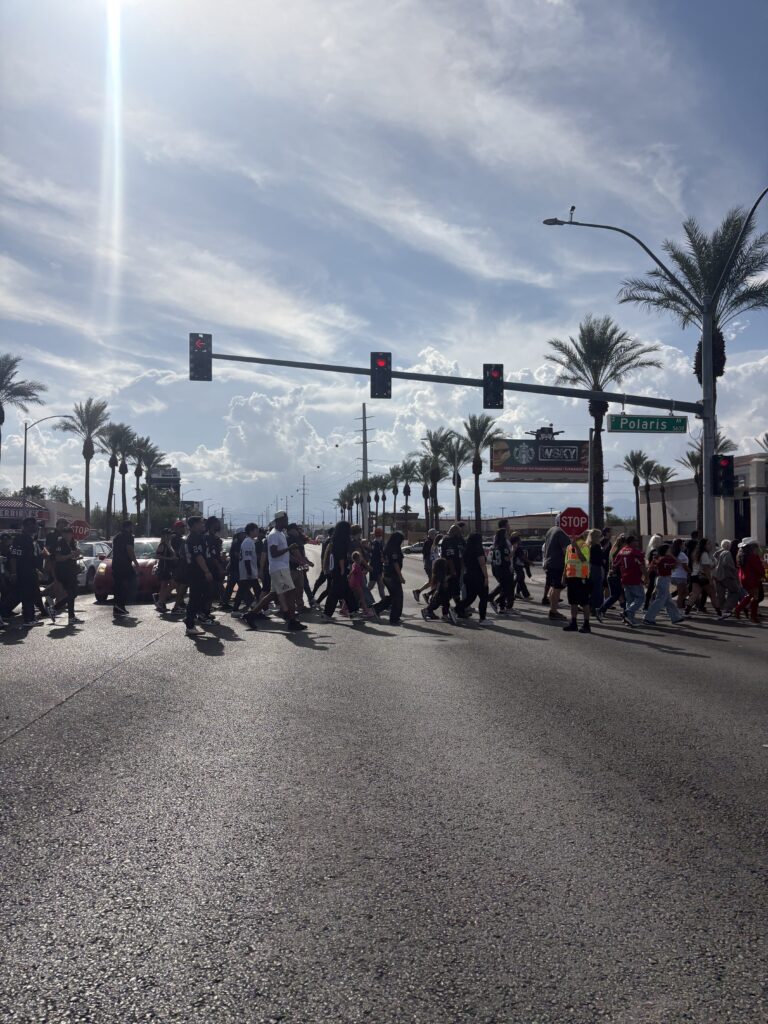 Crowd of Raiders fans crossing Polaris Avenue near Allegiant Stadium on game day, with a crossing guard directing traffic