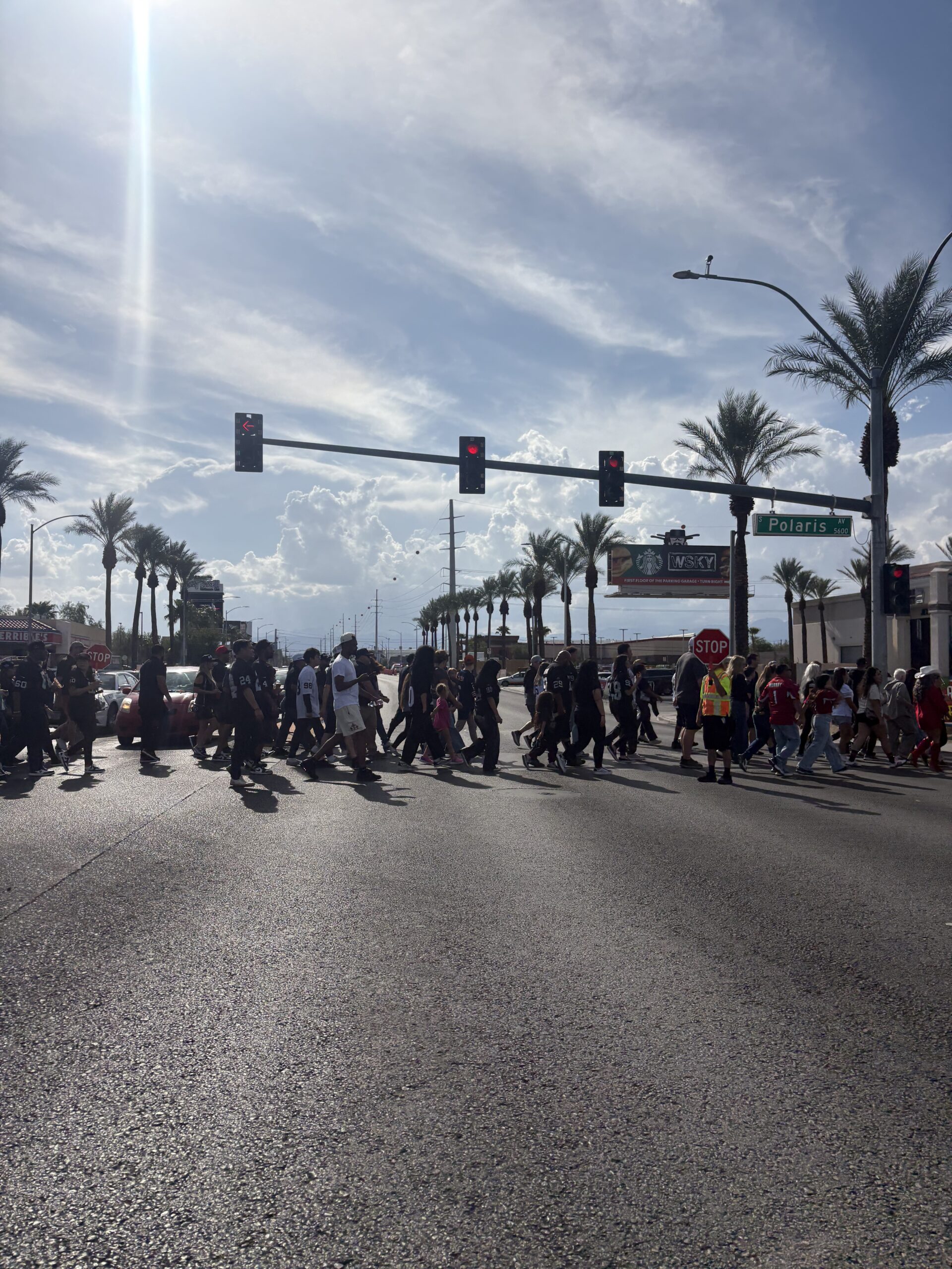 Crowd of Raiders fans crossing Polaris Avenue near Allegiant Stadium on game day, with a crossing guard directing traffic